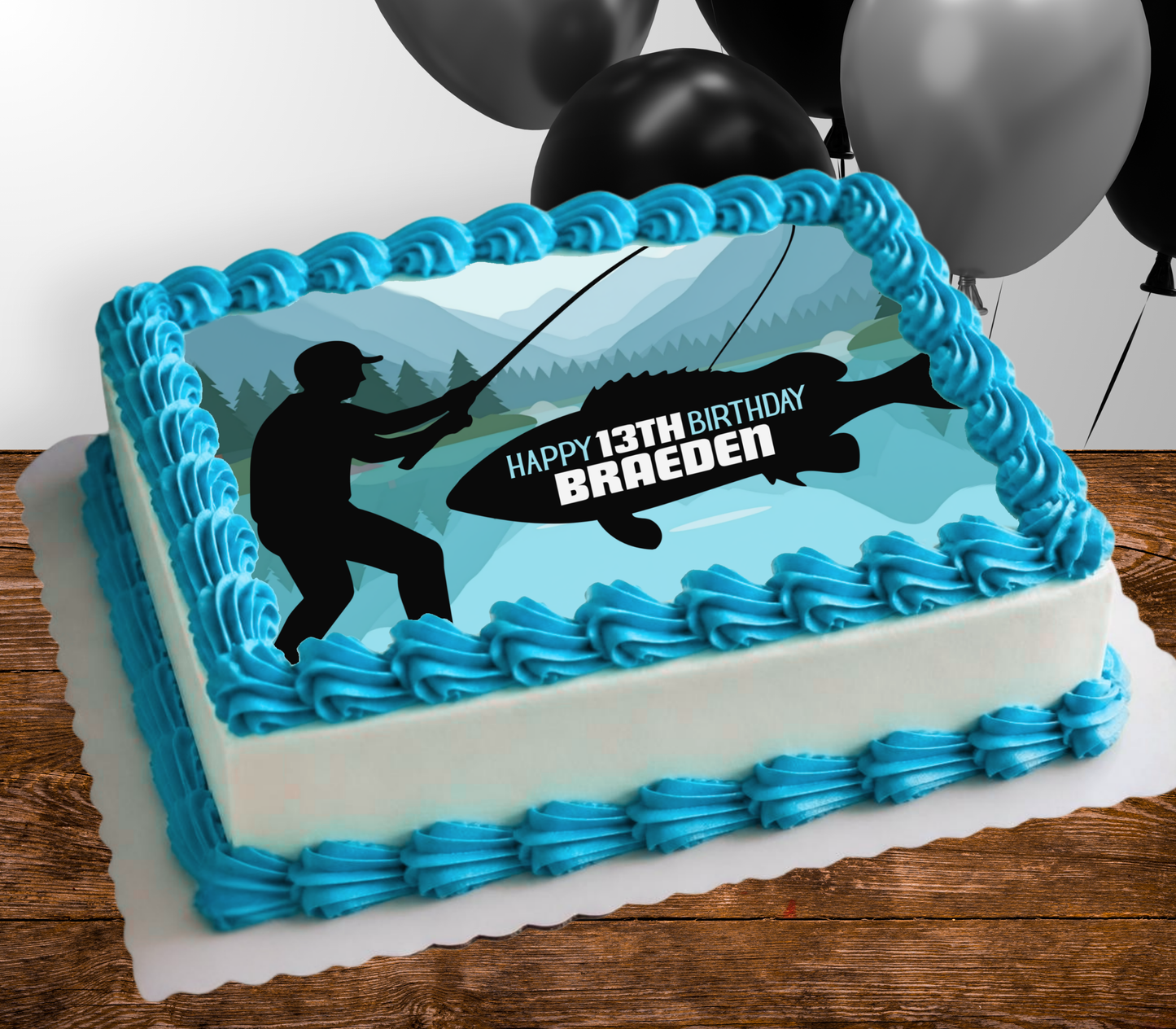 A rectangular birthday cake with blue and white frosting, decorated with a fishing-themed graphic showing a fisherman silhouette, a large fish, mountains, and the text 'HAPPY 13TH BIRTHDAY BRAEDEN'. Black and silver balloons are visible in the background against a white wall, with the cake sitting on a wooden surface.