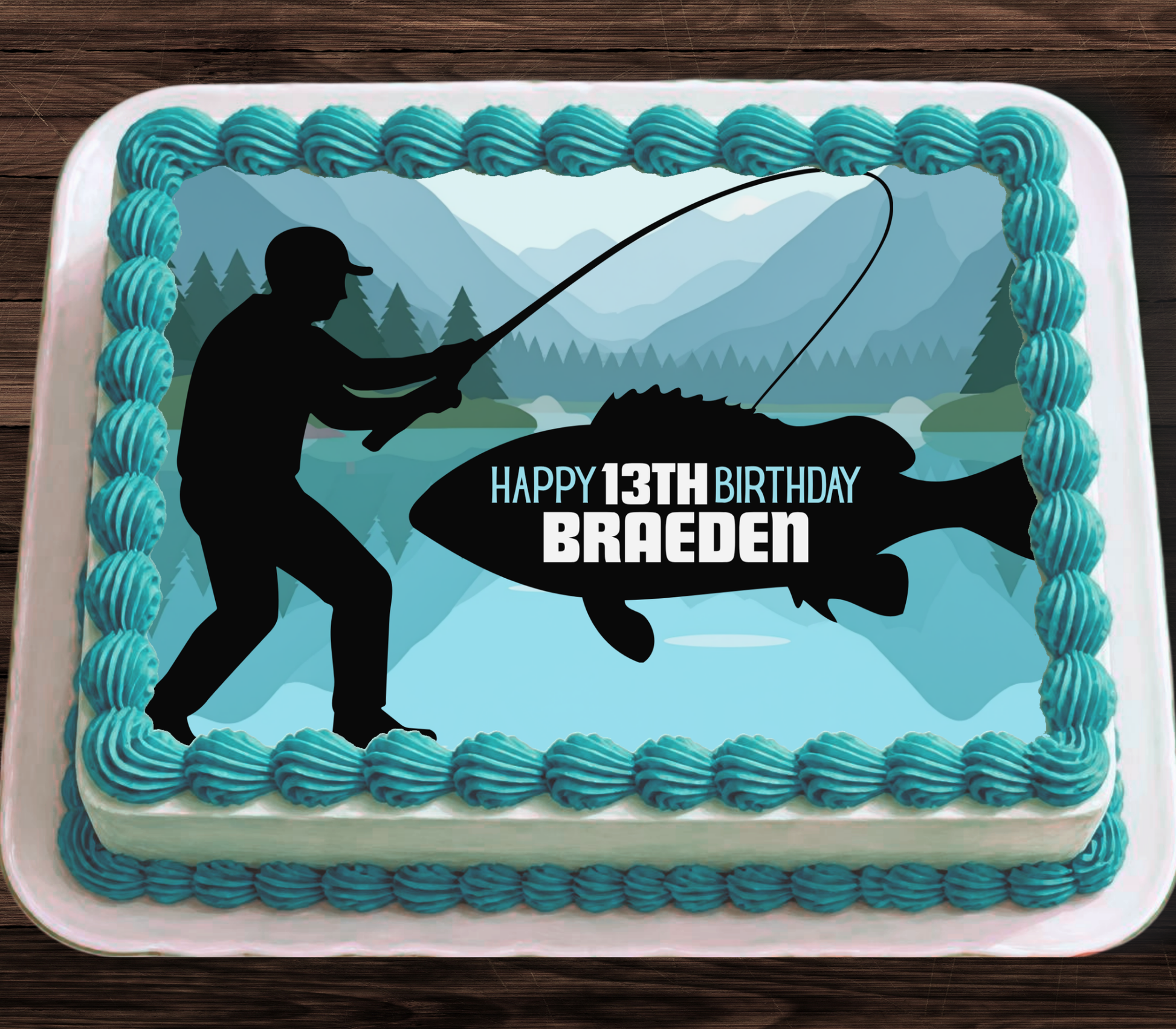 A rectangular cake decorated with turquoise frosting piped around the edges, featuring a fishing-themed graphic with a black silhouette of a fisherman, a large fish, mountains, and trees. The center reads 'HAPPY 13TH BIRTHDAY BRAEDEN' in white and black text.