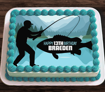 A rectangular cake decorated with turquoise frosting piped around the edges, featuring a fishing-themed graphic with a black silhouette of a fisherman, a large fish, mountains, and trees. The center reads 'HAPPY 13TH BIRTHDAY BRAEDEN' in white and black text.