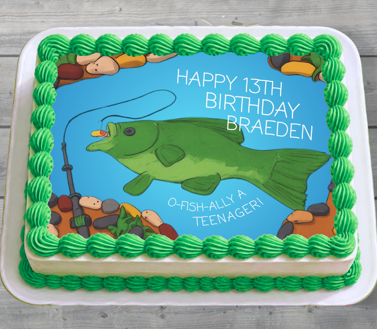 A square birthday cake decorated with green piped frosting around the border and a blue center panel showing a green fishing fish, rocks, and the text 'HAPPY 13TH BIRTHDAY BRAEDEN' and 'O-FISH-ALLY A TEENAGER!'.
