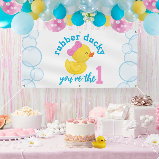 A baby first birthday party setup featuring a white banner with a yellow rubber duck, pink bow, and the text 'rubber ducky you've the 1'. The scene is decorated with pastel balloons, tinsel, a cake, wrapped gifts, and a small yellow rubber duck on a pink tablecloth.