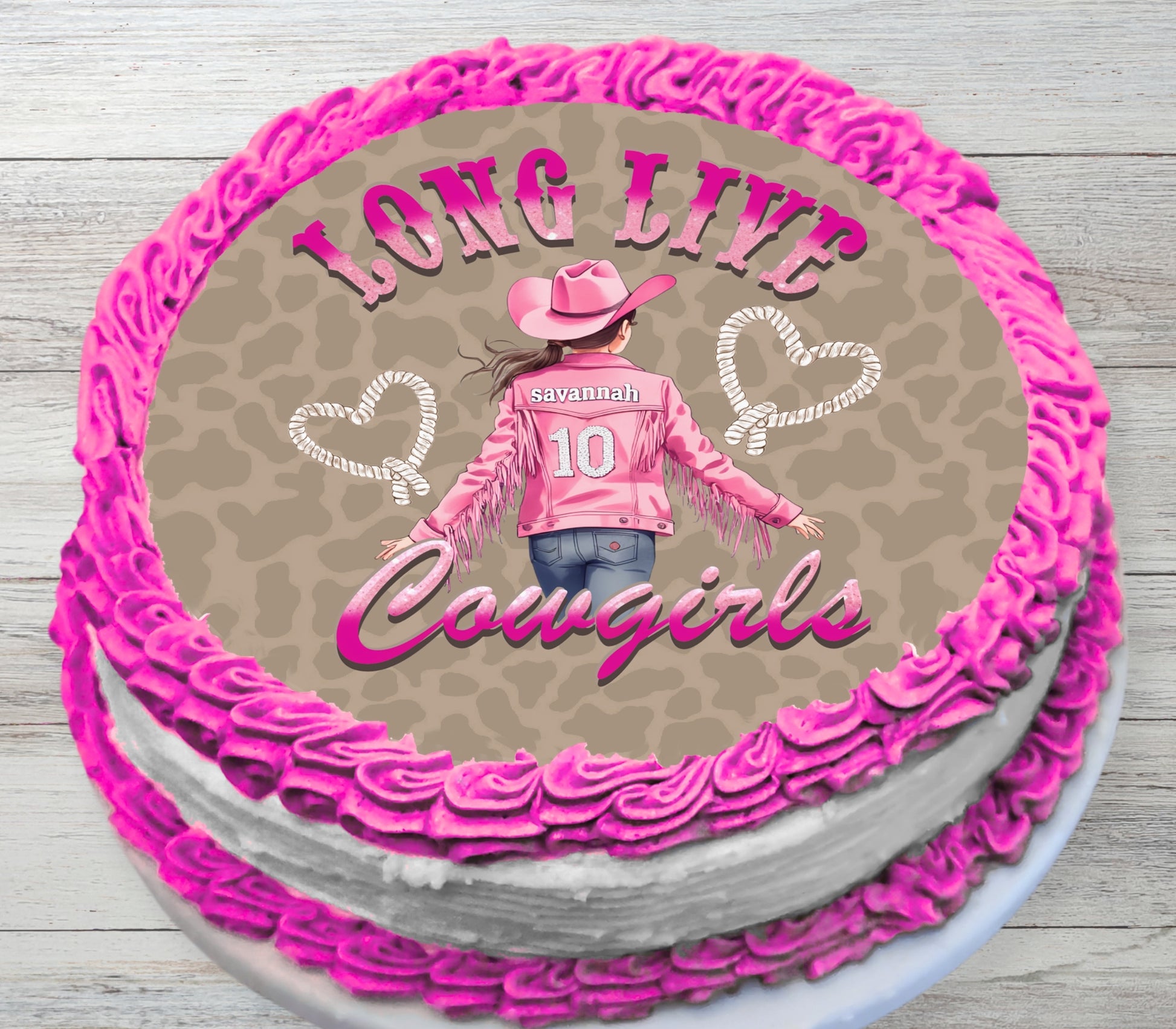 A round cake decorated with bright pink piped frosting around the edges and top, featuring a printed/custom graphic of a girl in a cowboy hat and jacket with the text 'LONG LIVE Cowgirls' on a tan patterned background.